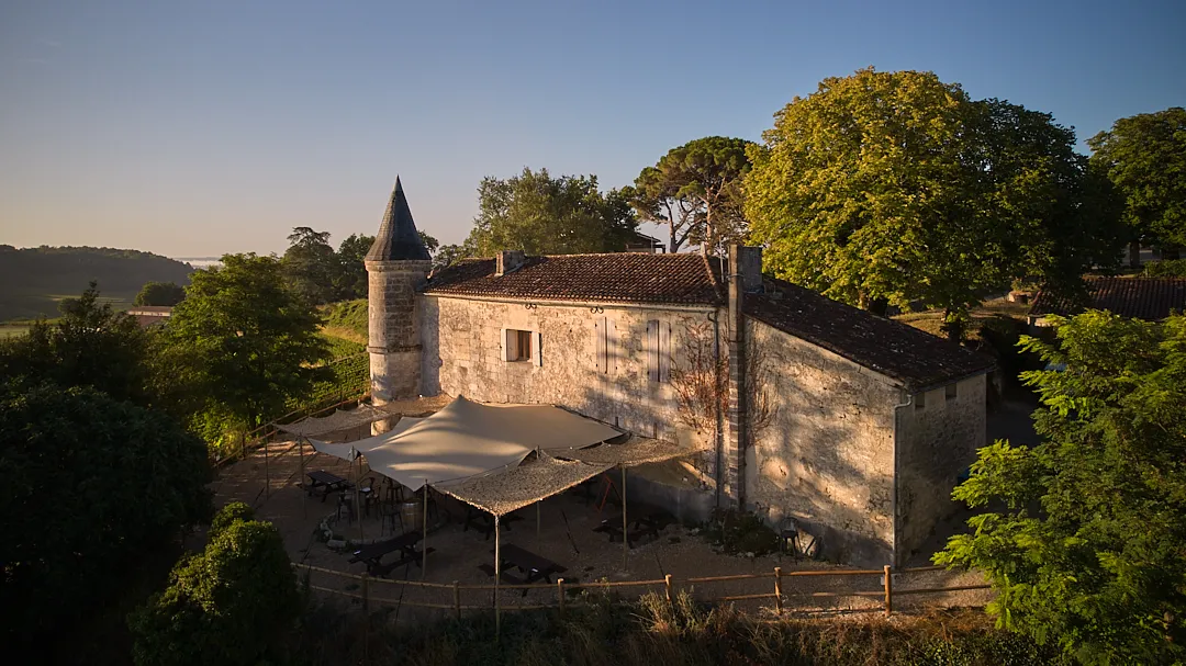 Aerial view of Château Tour Baladoz near Bordeaux, a stone building with a distinctive round tower, surrounded by green trees and featuring a shaded terrace wine bar