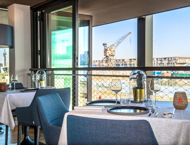Restaurant terrace with set tables, blue chairs, and panoramic view of Bordeaux port with industrial cranes in the background