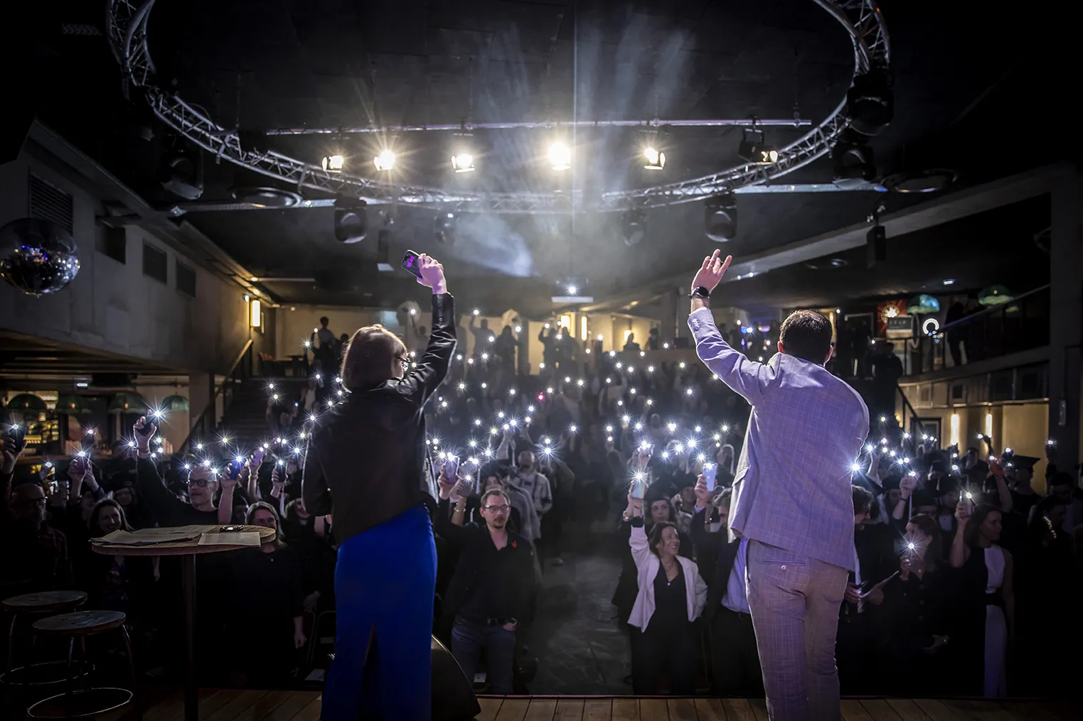 Two presenters raise their arms on stage before an audience holding illuminated phones during an indoor event in Bordeaux, under professional lighting fixtures and a lighting structure.