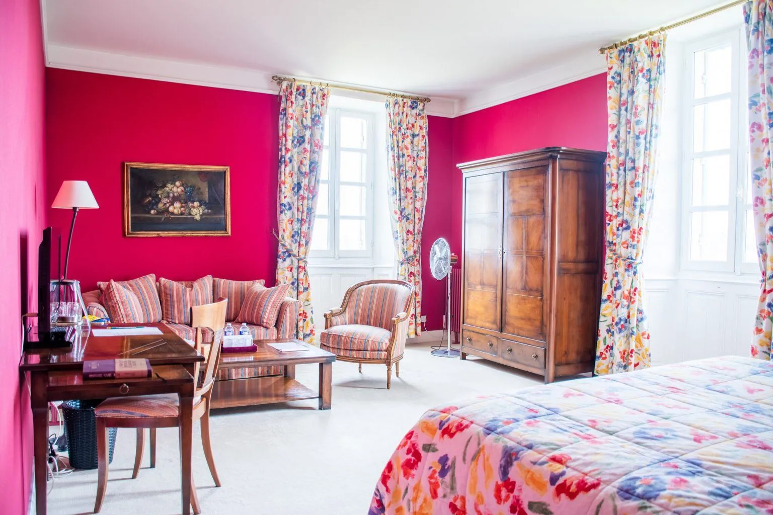 Luxury bedroom with vibrant pink walls, antique wooden furniture, colorful floral curtains, patterned floral bed and sitting area.