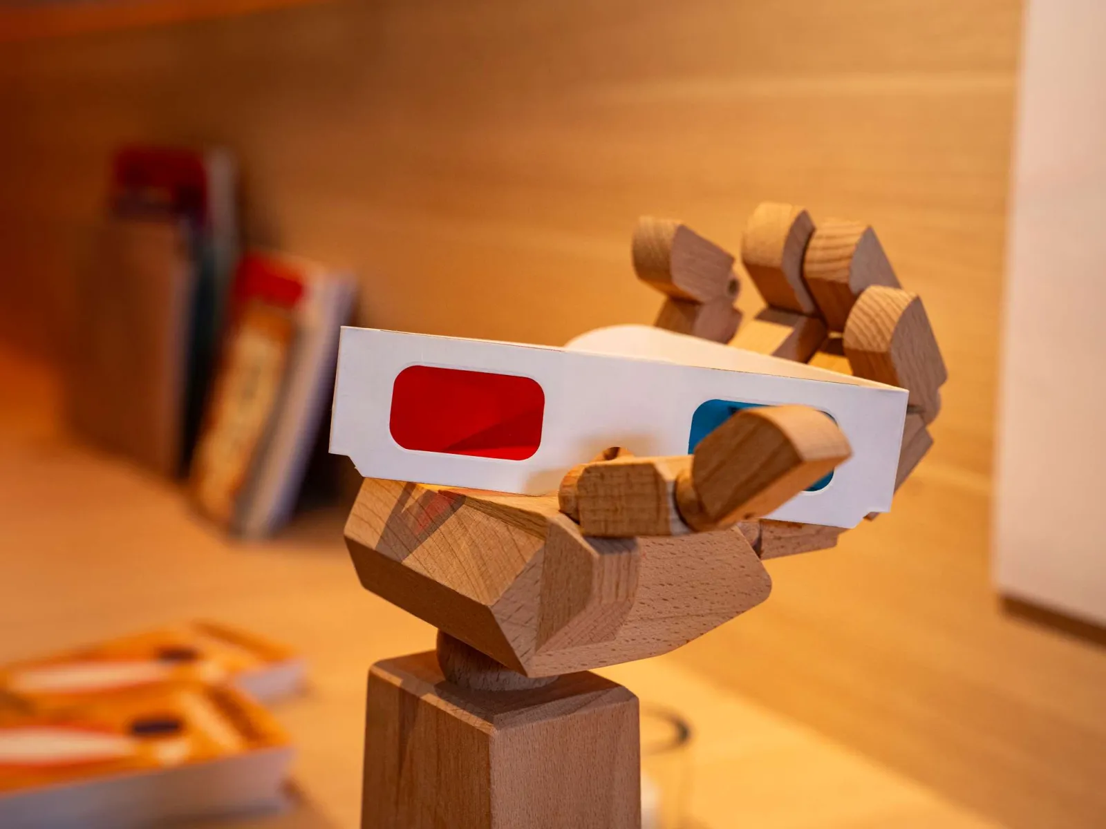 Wooden sculptural figure wearing red and white 3D glasses, mounted on a base in a warm, golden-toned interior setting.