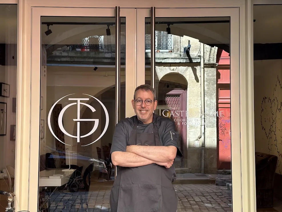 A chef smiles in front of The Gastronome Bordeaux storefront, a cooking school in Bordeaux, with arms crossed.