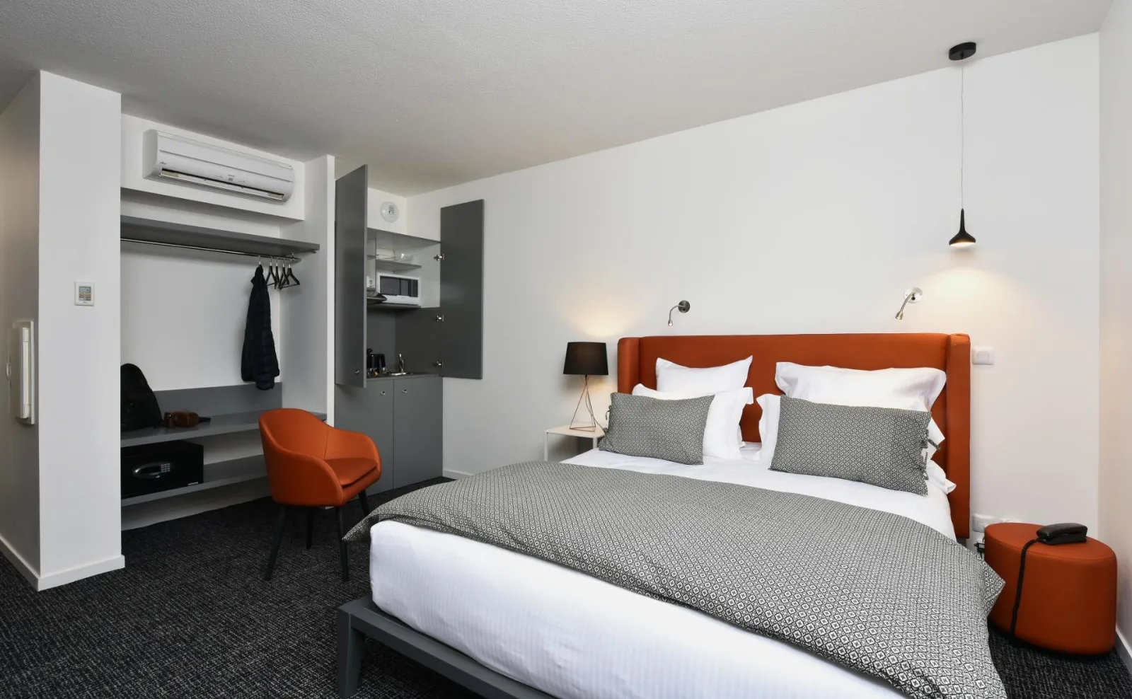 Modern hotel room at Ténéo Talence Espeleta in Bordeaux featuring a king-size bed with orange headboard, integrated work area, and gray and white furnishings.
