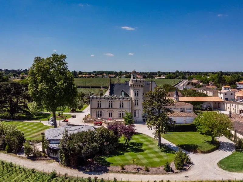 Aerial view of Château Pape Clément in Bordeaux, a white neo-gothic castle surrounded by formal gardens and vineyards, under a clear blue sky.