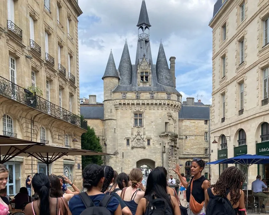 The Porte Cailhau gate in Bordeaux featuring distinctive stone towers with pointed roofs, framed by Haussmann-style buildings, with tourists photographing the scene