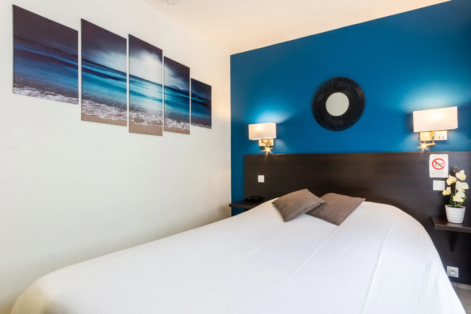 Modern hotel room with turquoise blue accent wall, white bed with gray pillows, black headboard, gold wall sconces, circular black mirror and multi-panel beach artwork. Hotel Notre Dame, Bordeaux.