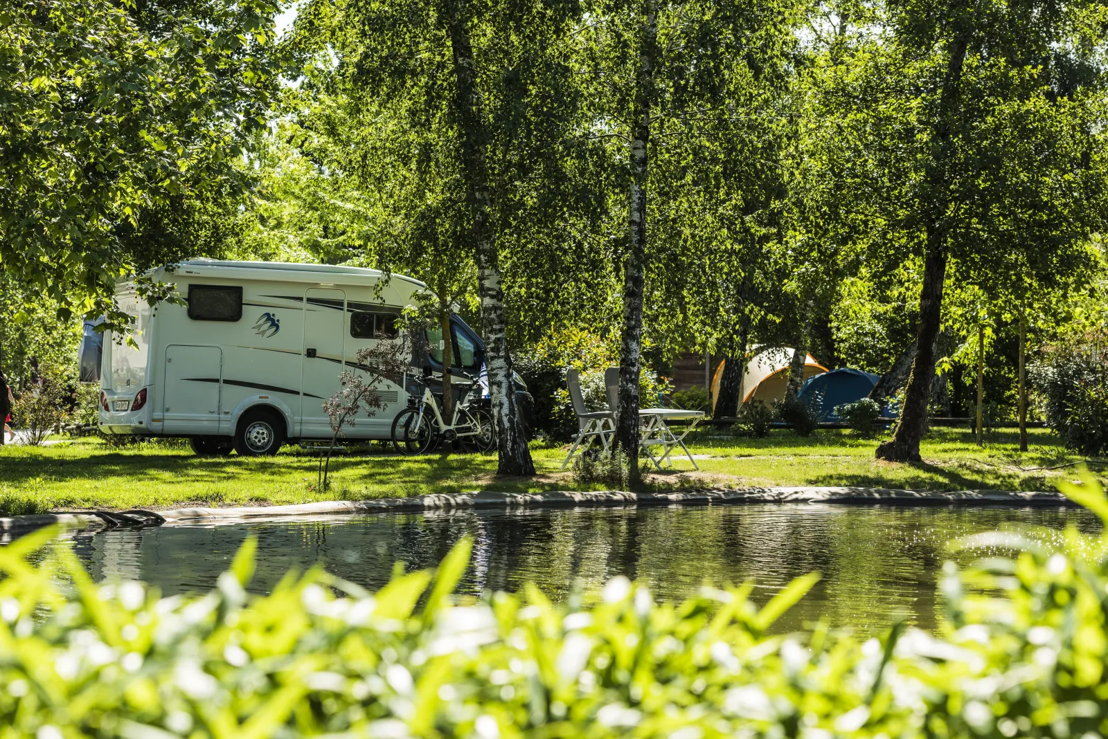 White motorhome parked by a lakeside surrounded by trees, with bicycles attached and a camping tent visible in the background at Yelloh! Village Bordeaux Lac.