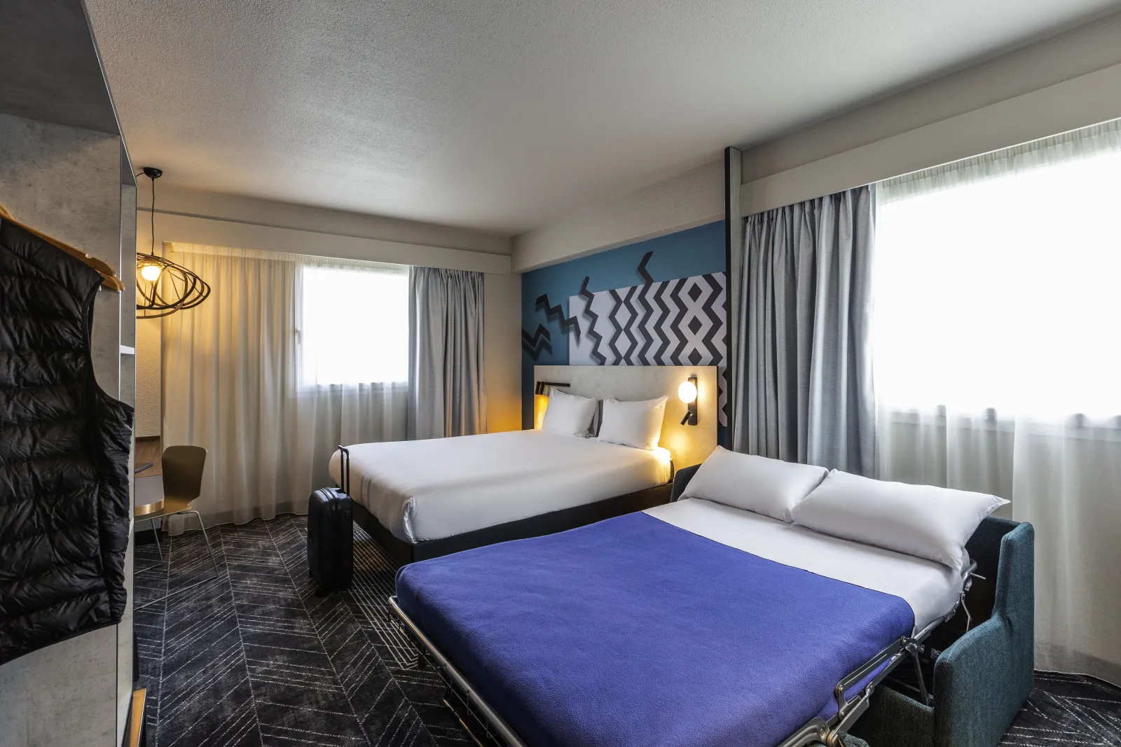 Modern hotel room with two beds (a blue double bed in the foreground and a white single bed in the background), geometric blue wall decoration, large windows with gray curtains, warm lighting and contemporary furniture.