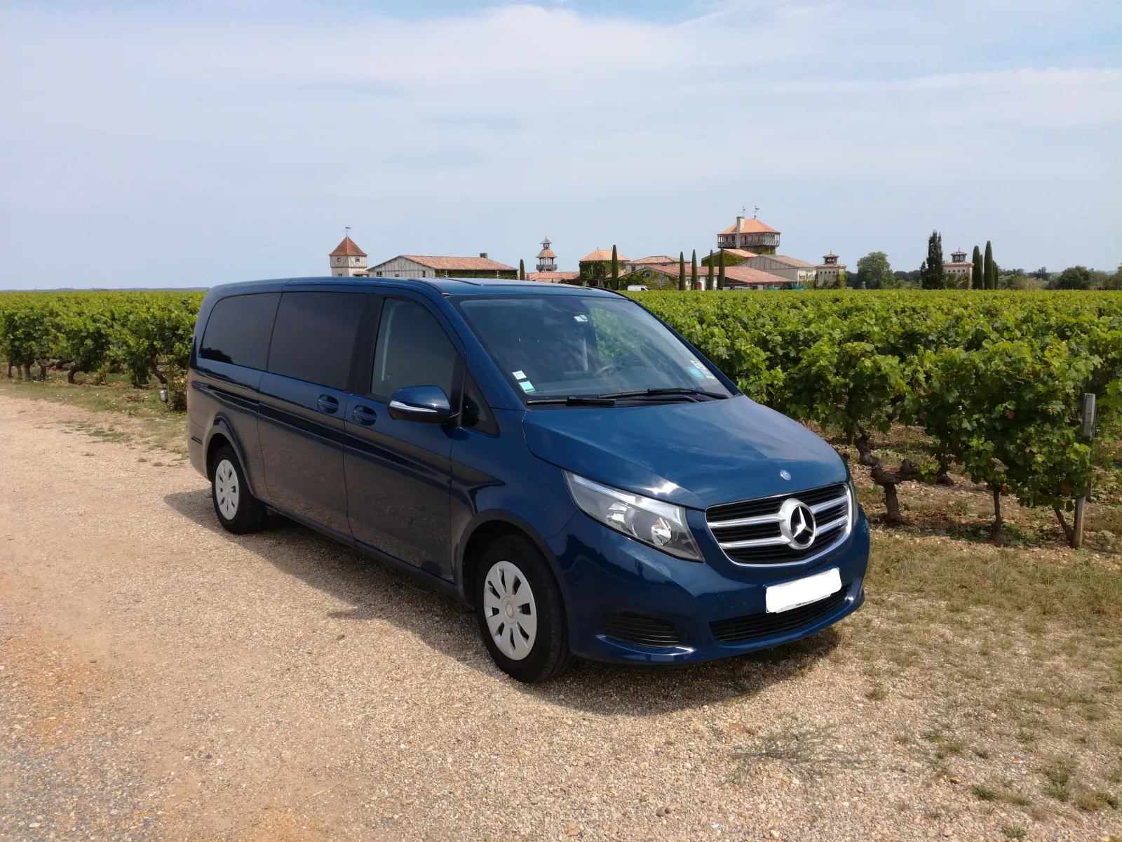Blue Mercedes minibus parked on a dirt road overlooking Bordeaux vineyards with a wine estate visible in the background.