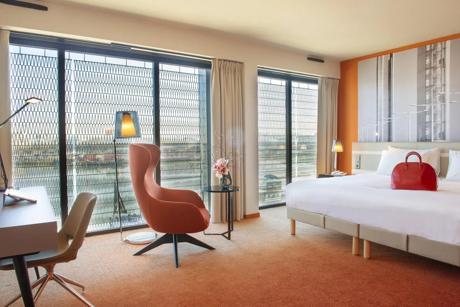 Spacious suite at Radisson Blu Hotel Bordeaux featuring two white beds, coral red armchair, floor-to-ceiling windows overlooking the city, and modern minimalist décor.