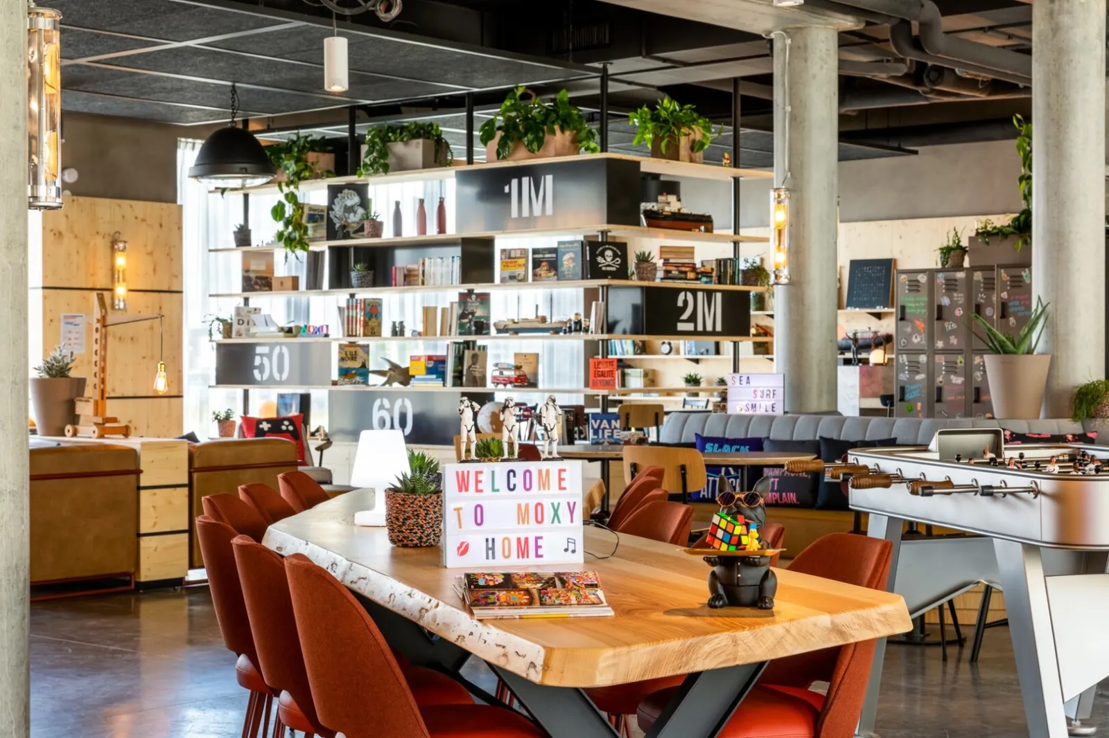 Modern interior of Moxy Bordeaux hotel featuring a welcoming common area with a wooden table, red chairs, white shelves decorated with green plants, a colorful welcome sign, and contemporary industrial ambiance.