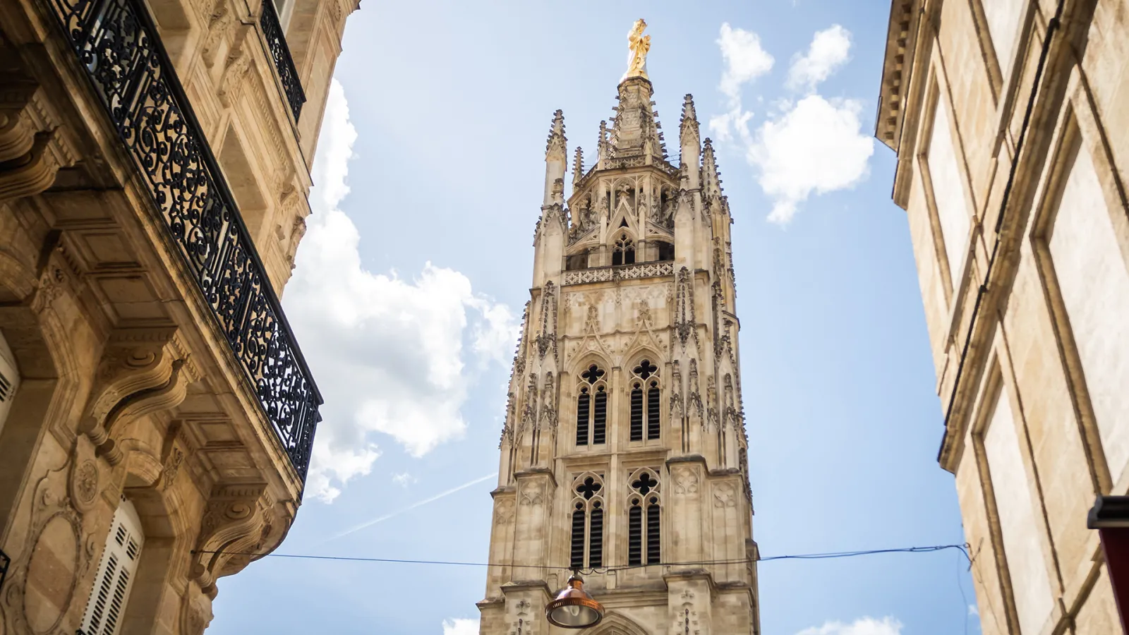 Pey-Berland Tower in Bordeaux, a Gothic stone tower with a golden spire, viewed from a narrow street between two Haussmann buildings.