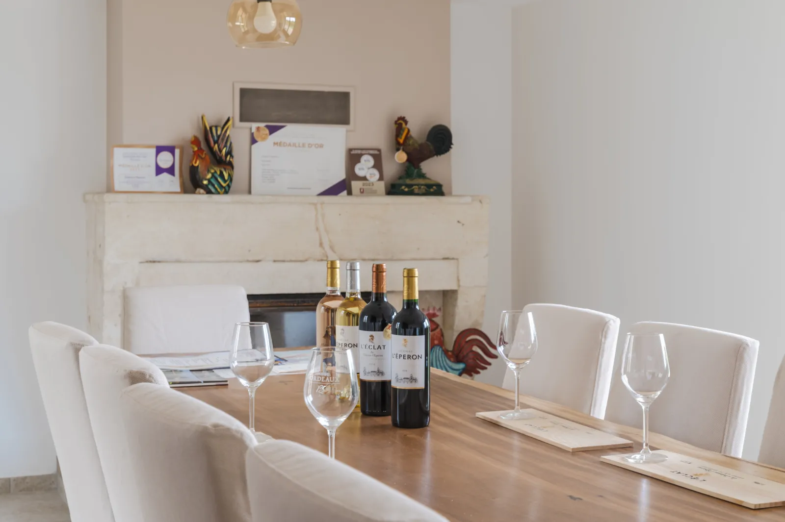 Tasting room at Château l'Éperon in Bordeaux featuring a wooden table with wine glasses and wine bottles, white chairs, and a fireplace decorated with trophies and medals in the background