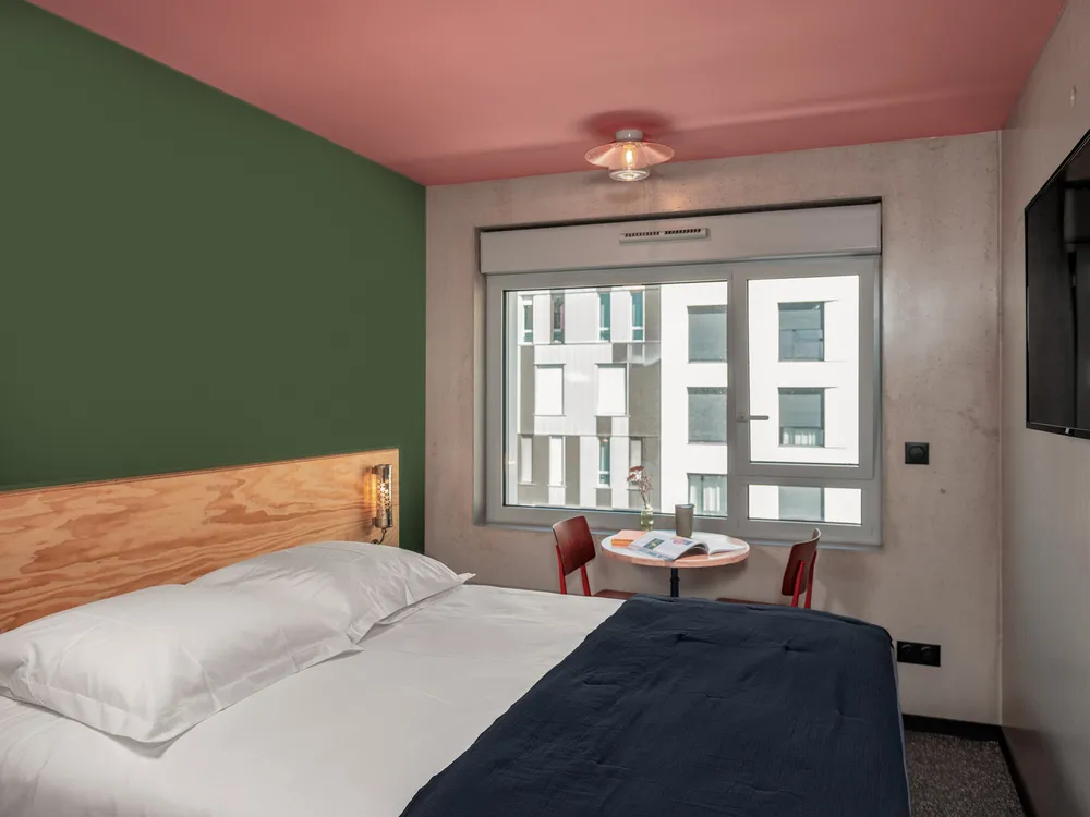 Modern hotel bedroom in Bordeaux featuring a green accent wall, pink ceiling, wooden bed frame, desk area, and window overlooking residential buildings