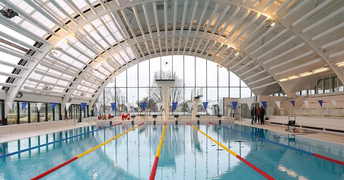 Interior of Bordeaux's Galin municipal swimming pool featuring an Olympic-sized basin with lane dividers in yellow and red ropes, beneath a modern curved glass roof.