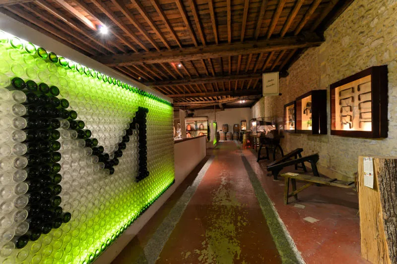 Interior of the Museum of Wine and Vine Crafts in Bordeaux, featuring an artistic installation of illuminated green and black bottles forming a graphic pattern on the left wall, with a terracotta exhibition corridor, display cases, and wooden ceiling beam