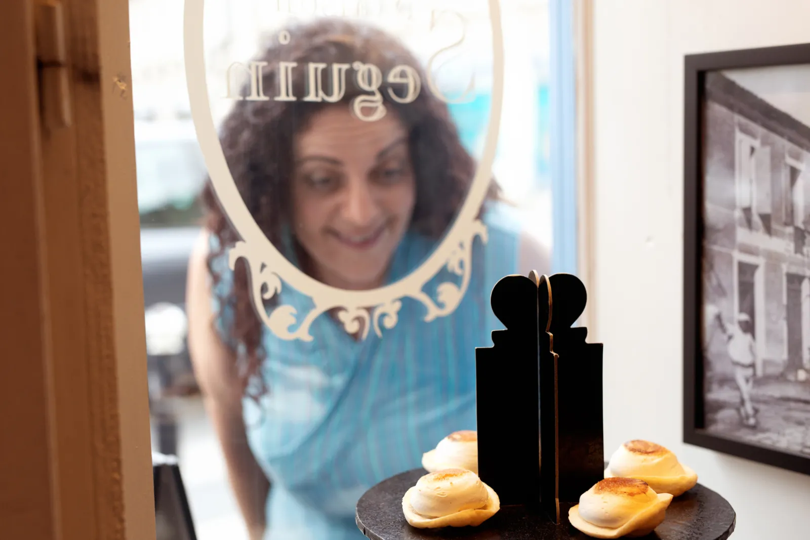 A smiling woman behind an ornate mirror gazes at golden pastries arranged in front of her in a Bordeaux interior.