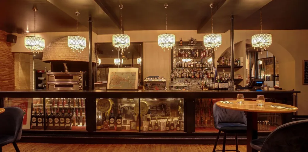 An elegant bar featuring a dark wooden counter, illuminated display cases with wine and spirits bottles, crystal pendant chandeliers, and modern seating in a warm, sophisticated setting.