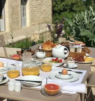 Elegant breakfast table set on a terrace with pastries, fresh juices, coffee, fruits and flowers, in front of a stone house with white shutters.