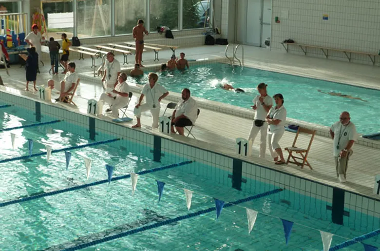 Swimming competition at the Olympic pool of Villenave d'Ornon near Bordeaux, with multiple swimmers in the water and judges seated along the pool deck.
