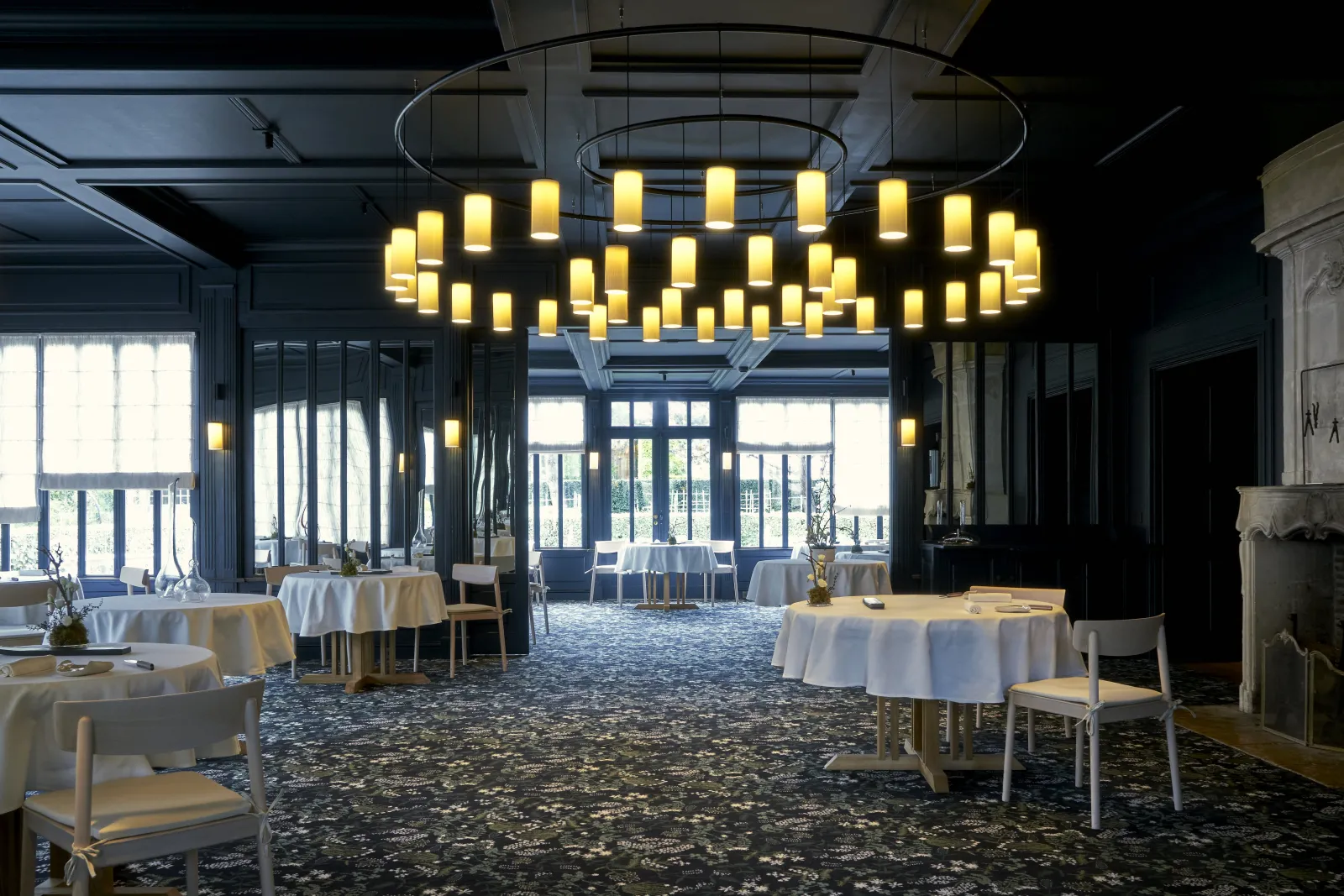 Elegant dining room at La Grand'Vigne in Bordeaux featuring round tables with white linens, modern brass chandelier, floor-to-ceiling windows, and contemporary minimalist decor.