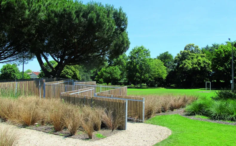 Public park along the Jalle de Gajac waterfront in Bordeaux featuring green lawns, ornamental grasses, wooden and metal fencing, mature trees and clear sky