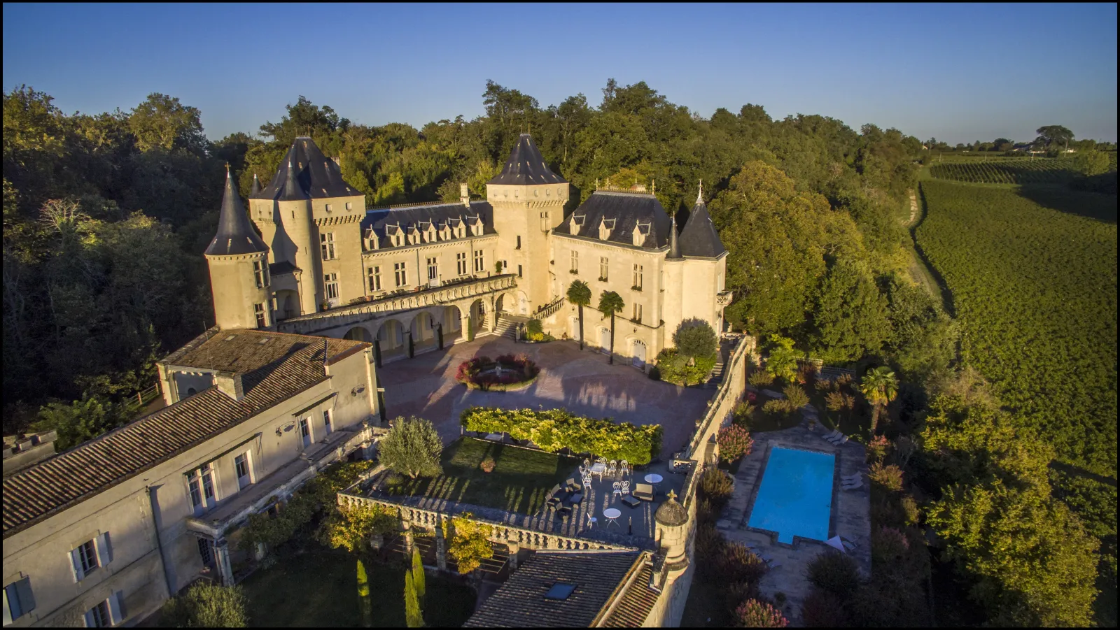 Aerial view of Château de La Rivière near Bordeaux, featuring Renaissance architecture with conical towers, paved courtyard, swimming pool, and surrounding vineyards.