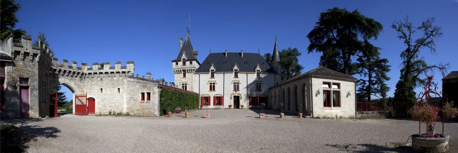 Inner courtyard of Château de Pressac in Bordeaux featuring white stone buildings with red doors, crenellated towers and pointed roofs against a clear blue sky.