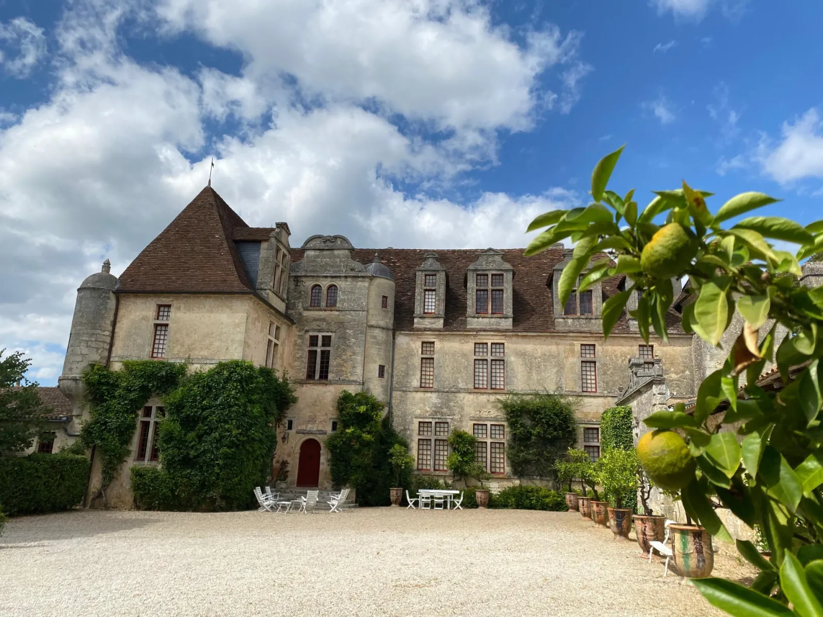 Frontal view of Château Le Grand Verdus in Bordeaux, a stone manor house with a tower, surrounded by greenery and grapevines, under a partly cloudy blue sky.