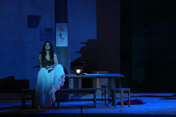 Scene from the opera Carmen at Bordeaux National Theatre: a woman in traditional costume sitting at an illuminated table under blue stage lighting, with minimalist set design and stairs in the background.