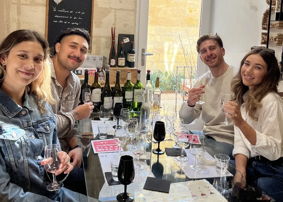 Four smiling people enjoy a wine tasting around a table with multiple bottles of Bordeaux wine and glasses in a wine cellar or tasting room in Bordeaux.