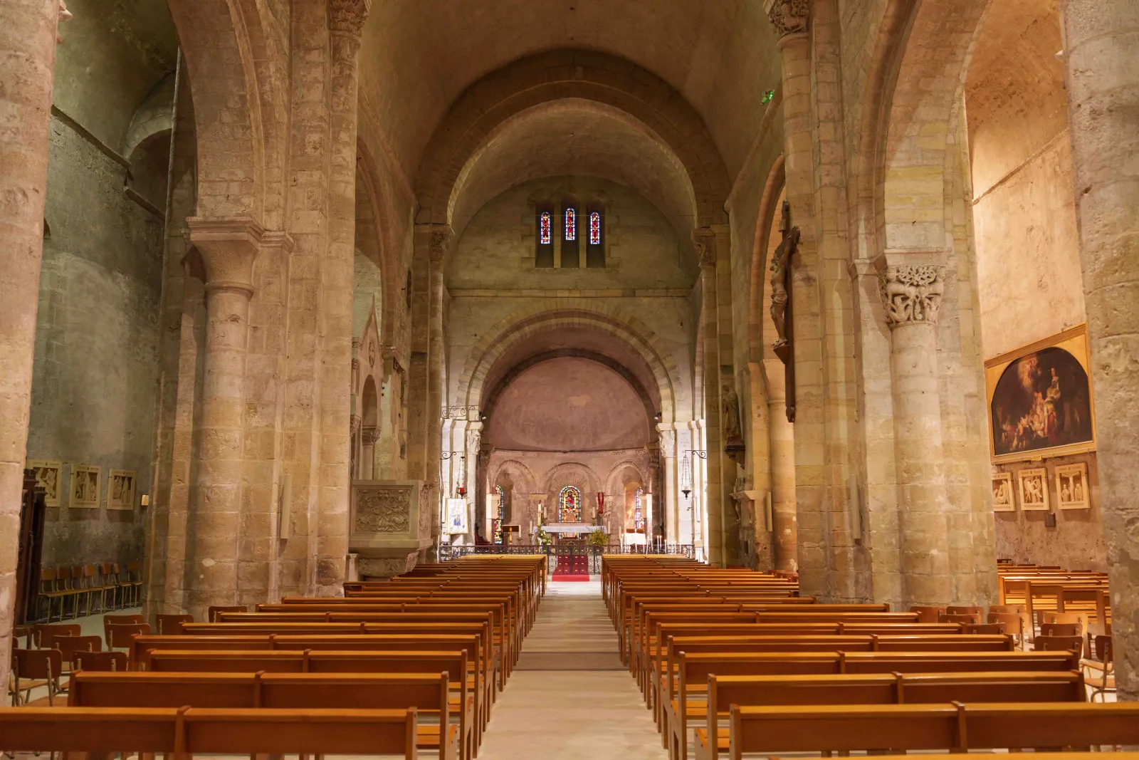 Interior of the Basilica of Notre-Dame-de-la-fin-des-Terres in Bordeaux, featuring the nave with Romanesque columns, barrel vaults, wooden pews, and the altar at the far end lit by stained glass windows.