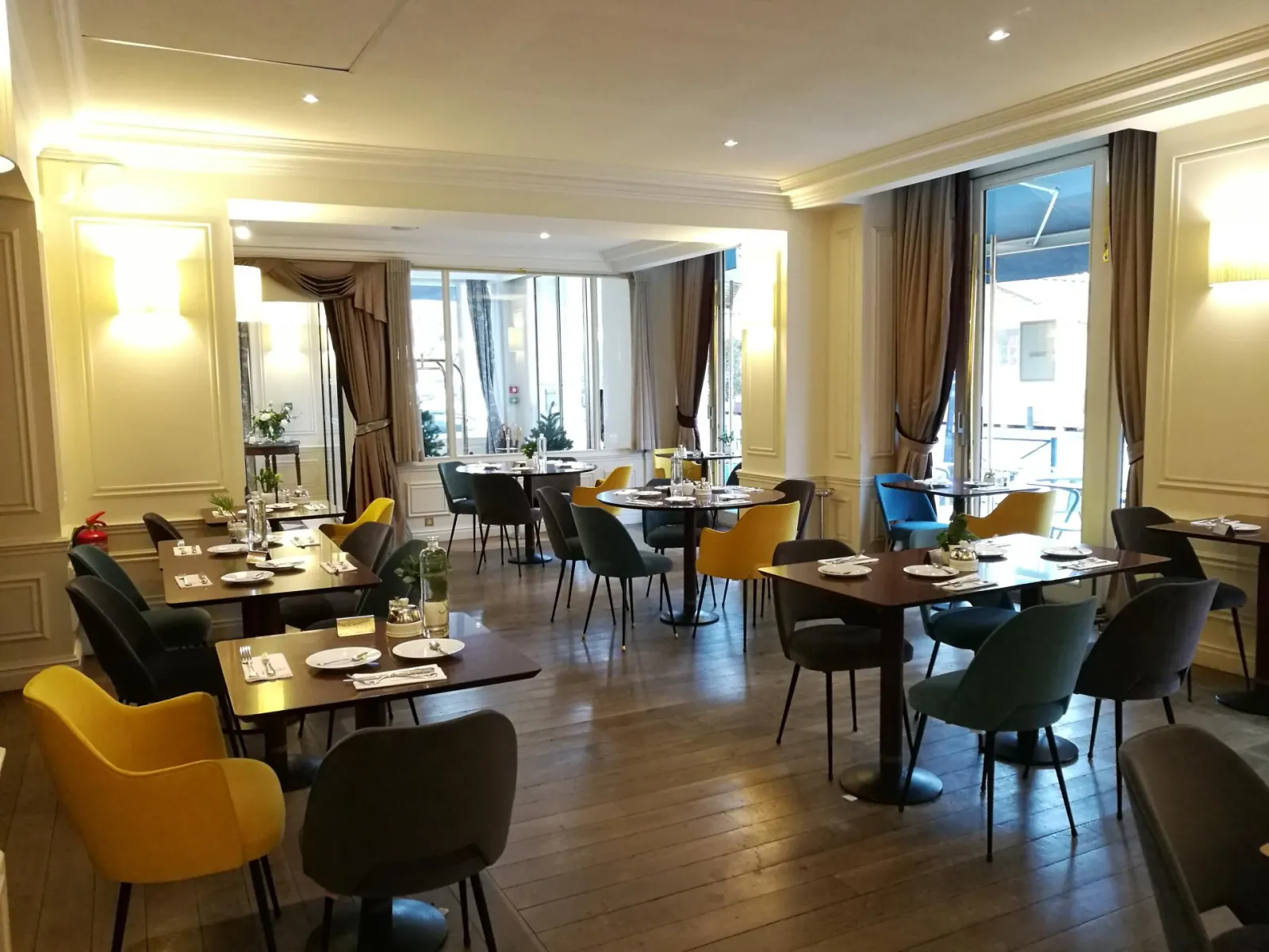 Elegant dining room at Comptoir de Sèze in Bordeaux featuring black tables, yellow and blue chairs, large windows, beige and gold decor, with an outdoor terrace view.