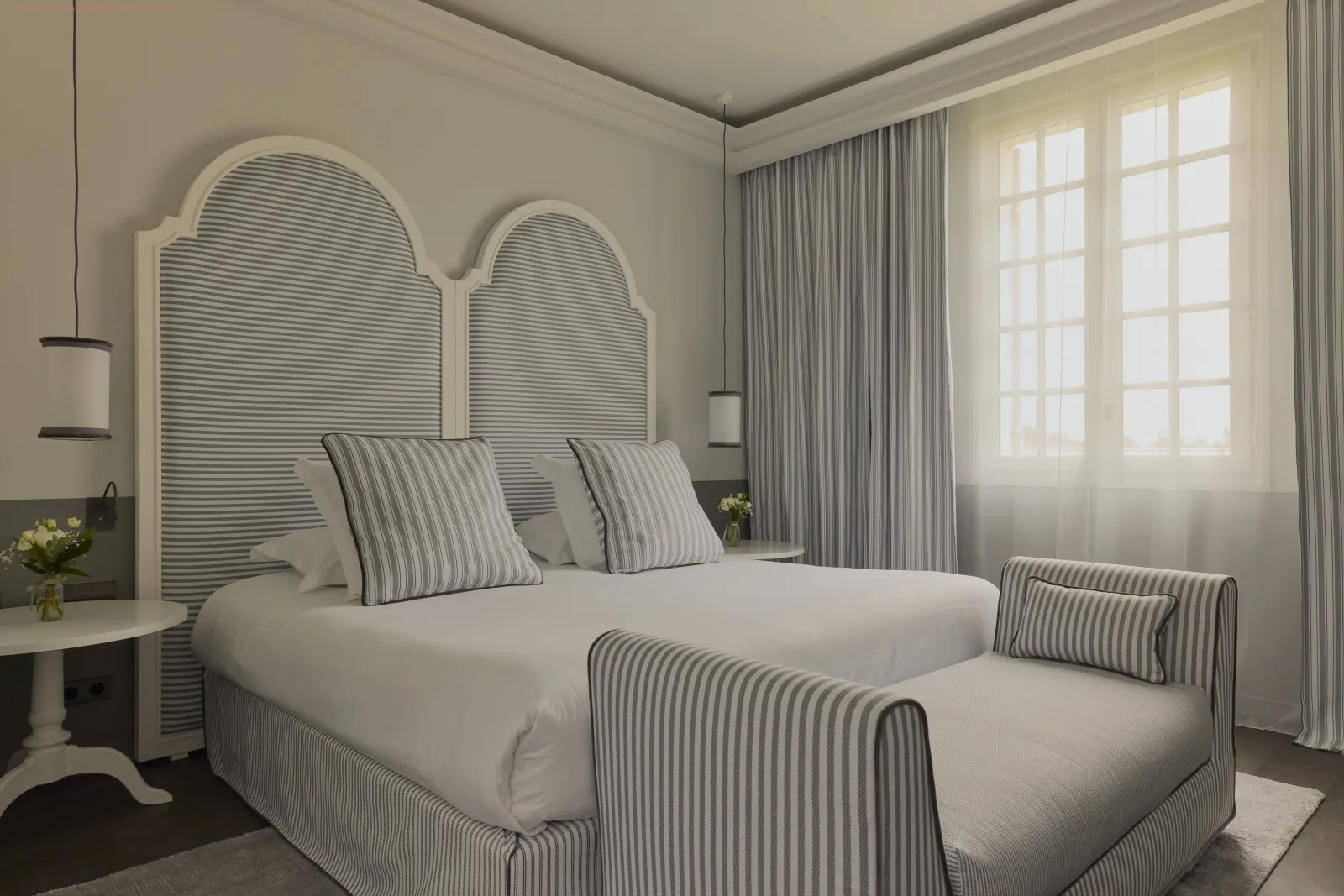 Elegant hotel bedroom featuring a striped grey and white double bed with matching arched headboard, striped pillows, large windows with grey curtains, white nightstand and pendant lights.