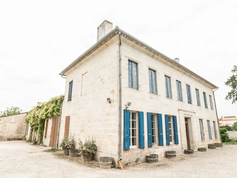 19th-century manor house with white stone facade, blue shutters and chimneys, surrounded by vineyards near Bordeaux