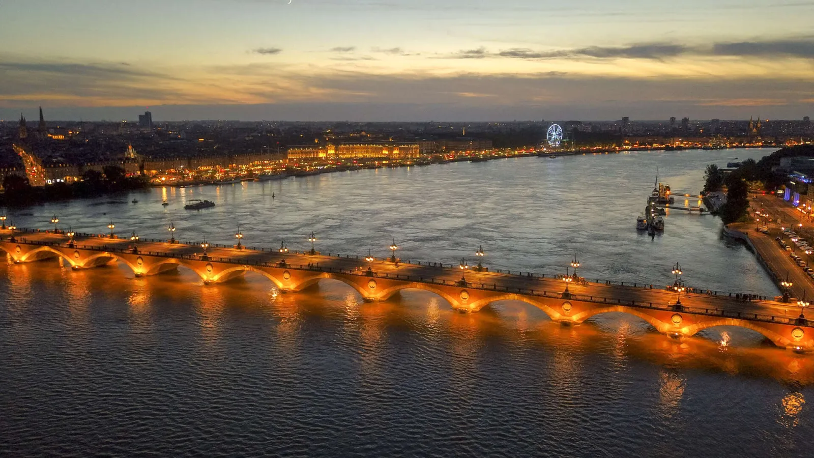 Twilight view of the illuminated Pont de Pierre in Bordeaux spanning the Garonne River with the city skyline in the background.
