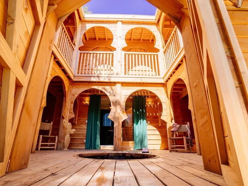 Wooden interior of a building featuring two-level gallery, Moorish arches, turquoise doors, and central courtyard open to blue sky at EcÔtelia estate in Bordeaux.