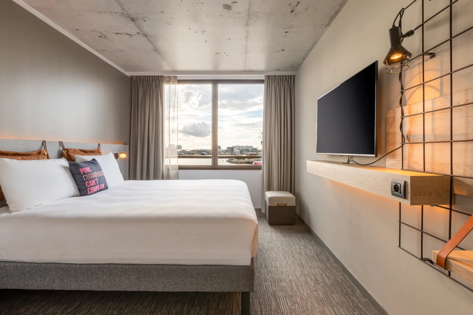 Modern hotel room at Moxy Bordeaux featuring a white bed, wall-mounted TV, large windows overlooking Bordeaux cityscape, and contemporary décor in neutral tones and wood accents.