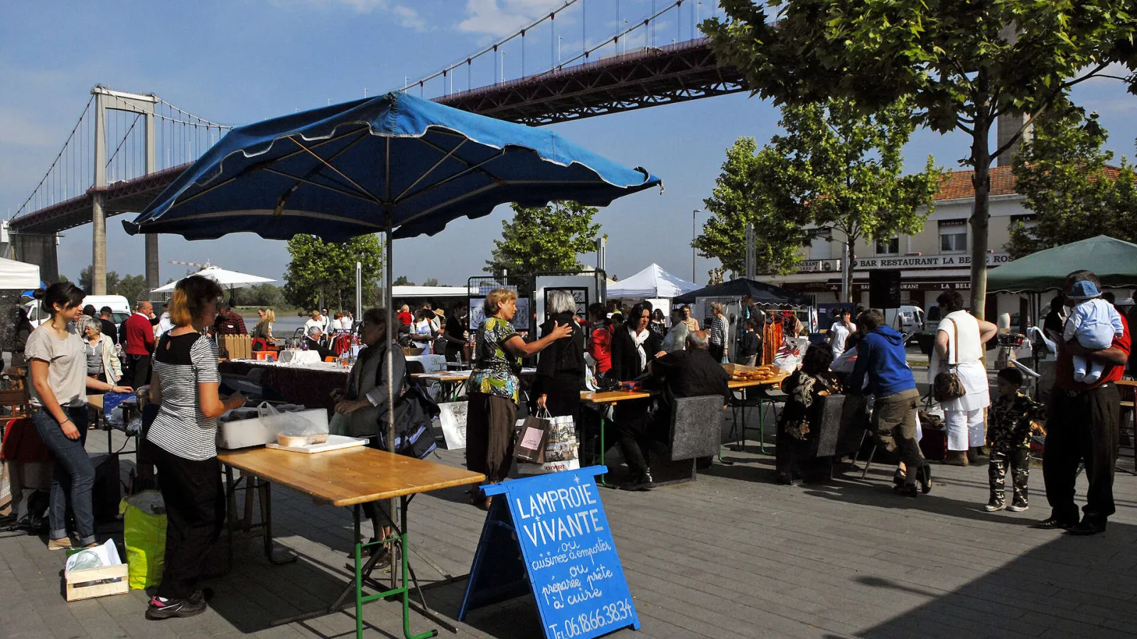 Outdoor market in Lormont with visitors under blue umbrellas, product stalls, and the Aquitaine Bridge visible in the background.