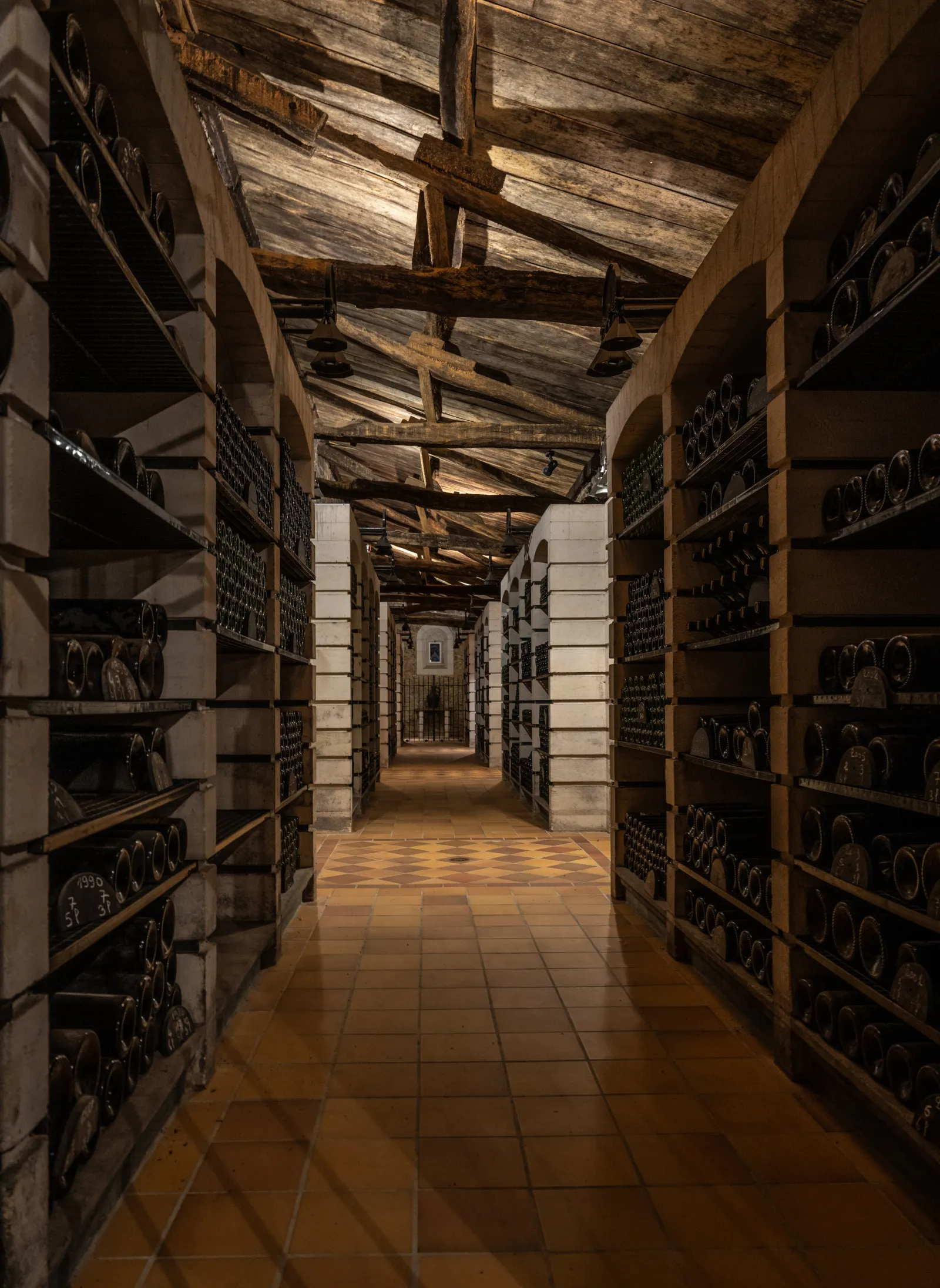 Underground wine cellar corridor at Château Carbonnieux in Bordeaux featuring wine bottle racks on both sides, exposed wooden ceiling beams, and tiled flooring.