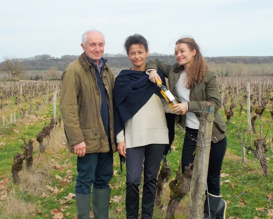 Three people pose in a vineyard at Château du Mayne La Merci near Bordeaux, holding a wine bottle, with rows of vines in the background.