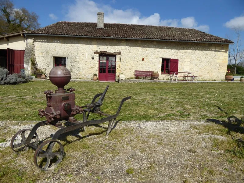 Facade of Château de Lisennes near Bordeaux featuring red shutters, light stone walls and antique wrought iron farm equipment in the foreground