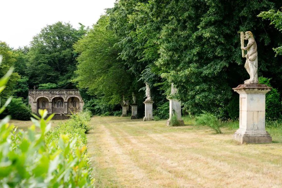 Tree-lined pathway at Château de Malle in Bordeaux featuring stone statues on pedestals, historic building in background, surrounded by lush greenery.