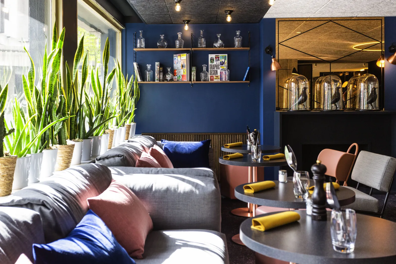Modern hotel room at Mercure Bordeaux Centre featuring blue walls, gray bed with colorful pillows, green plants by the window, shelves displaying glassware, and round work tables.