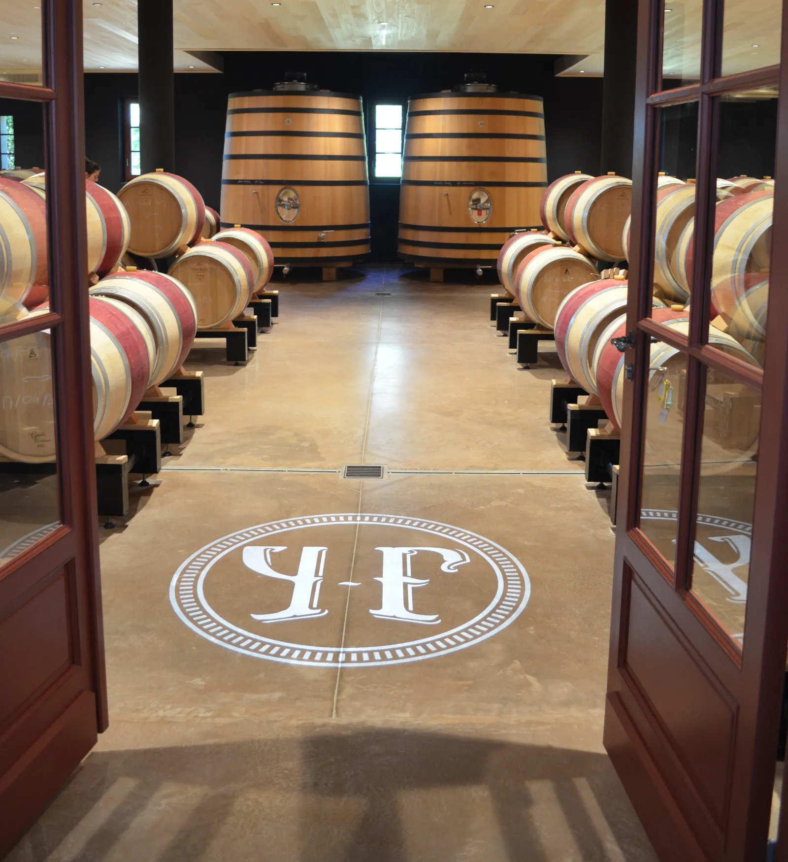 Interior of a wine cellar at Château Yon-Figeac in Bordeaux, featuring rows of wooden barrels and two large central vats, with the YF logo displayed on the marble floor.