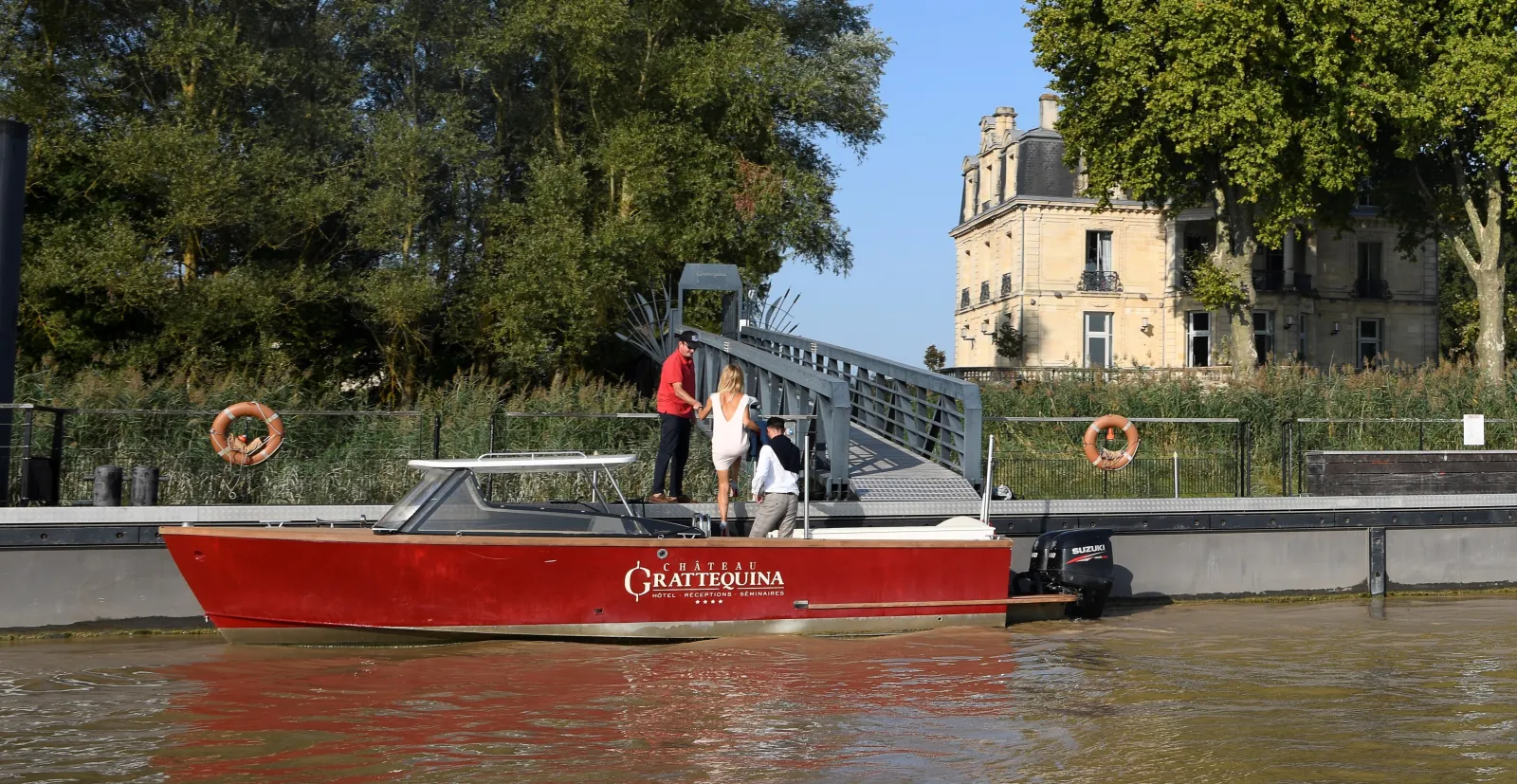 Red tourist boat Château Grattequina sailing on the Garonne River in Bordeaux, with passengers aboard and a historic château in the background