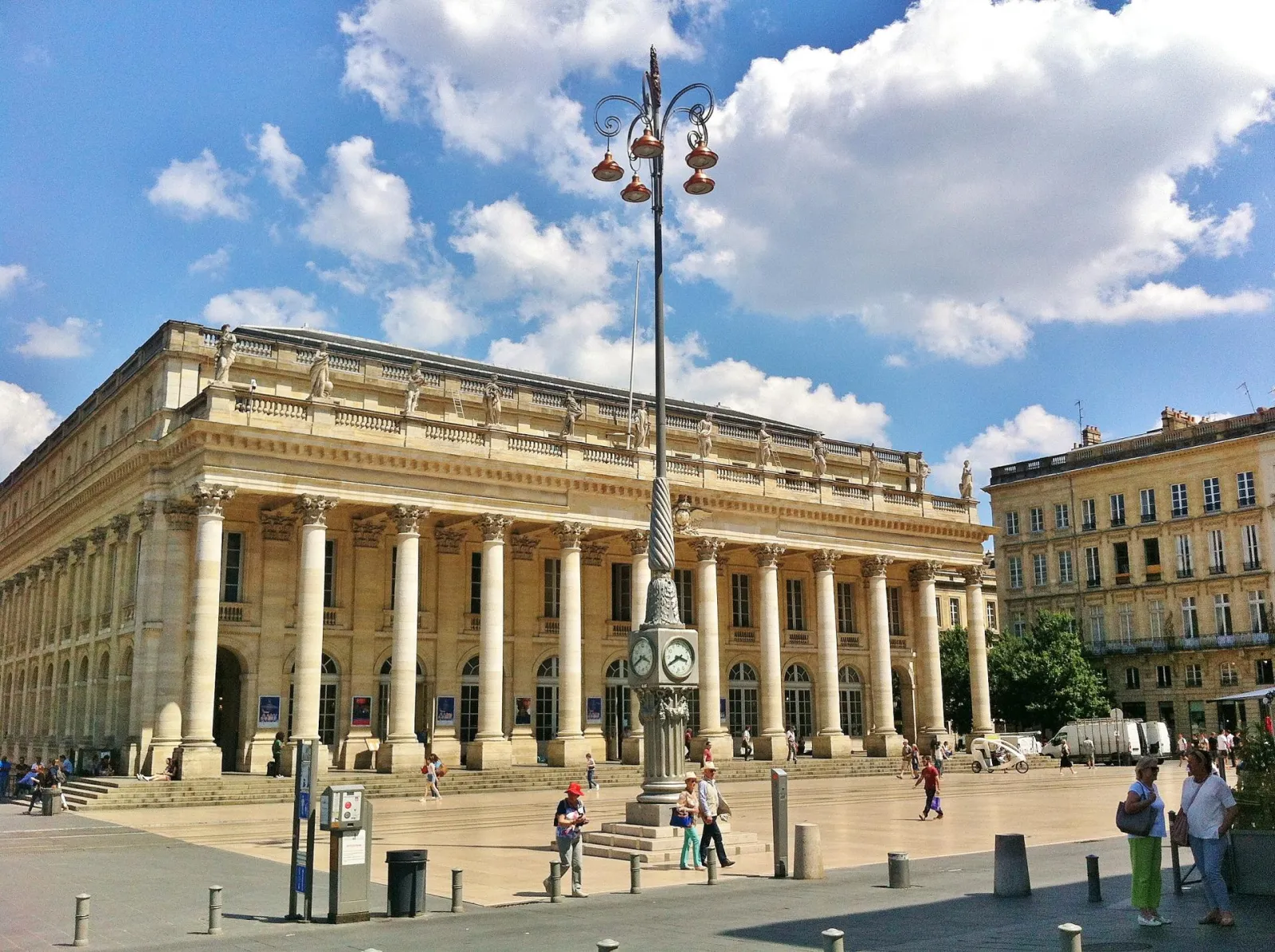 The Grand Theatre of Bordeaux, a striking neoclassical building with white columns on the Place de la Comédie, featuring a paved esplanade and ornate street lamp under a partly cloudy blue sky.