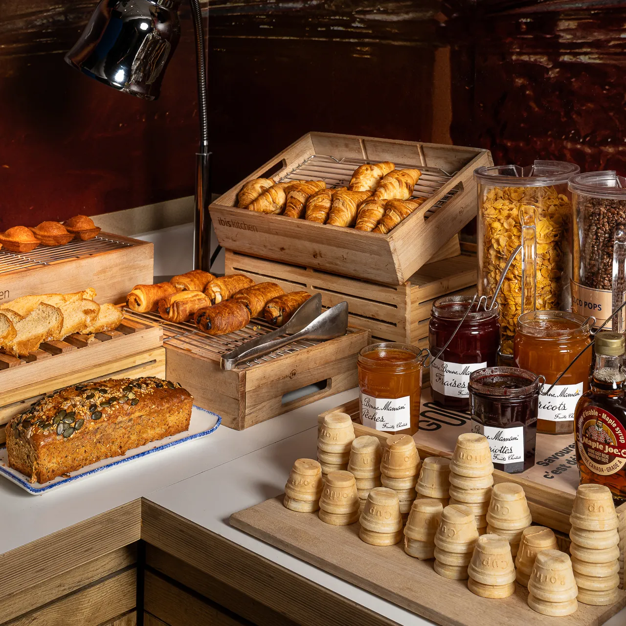 French breakfast buffet display featuring croissants, toasted bread, jam jars, cereals, and fruit juices under warm lighting