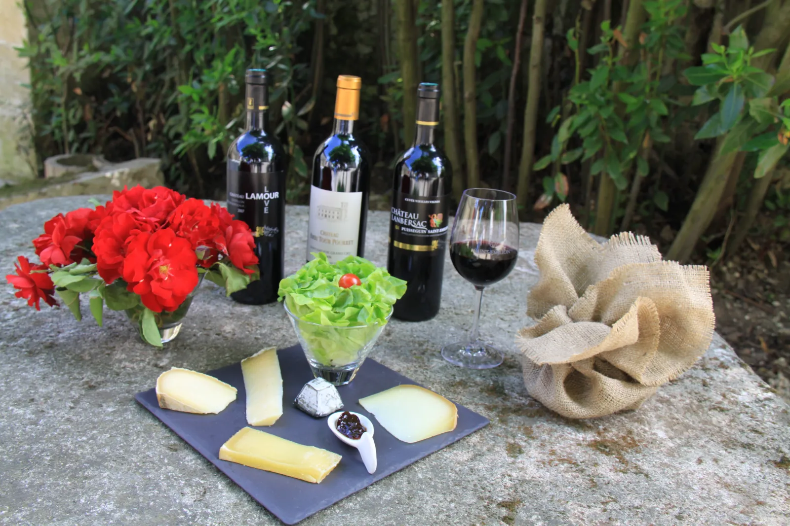 Wine and cheese tasting in a Bordeaux garden: three red wine bottles, a filled glass, cheeses on slate, red flowers, fresh lettuce and beige linen napkin, with ivy-covered wall in the background.
