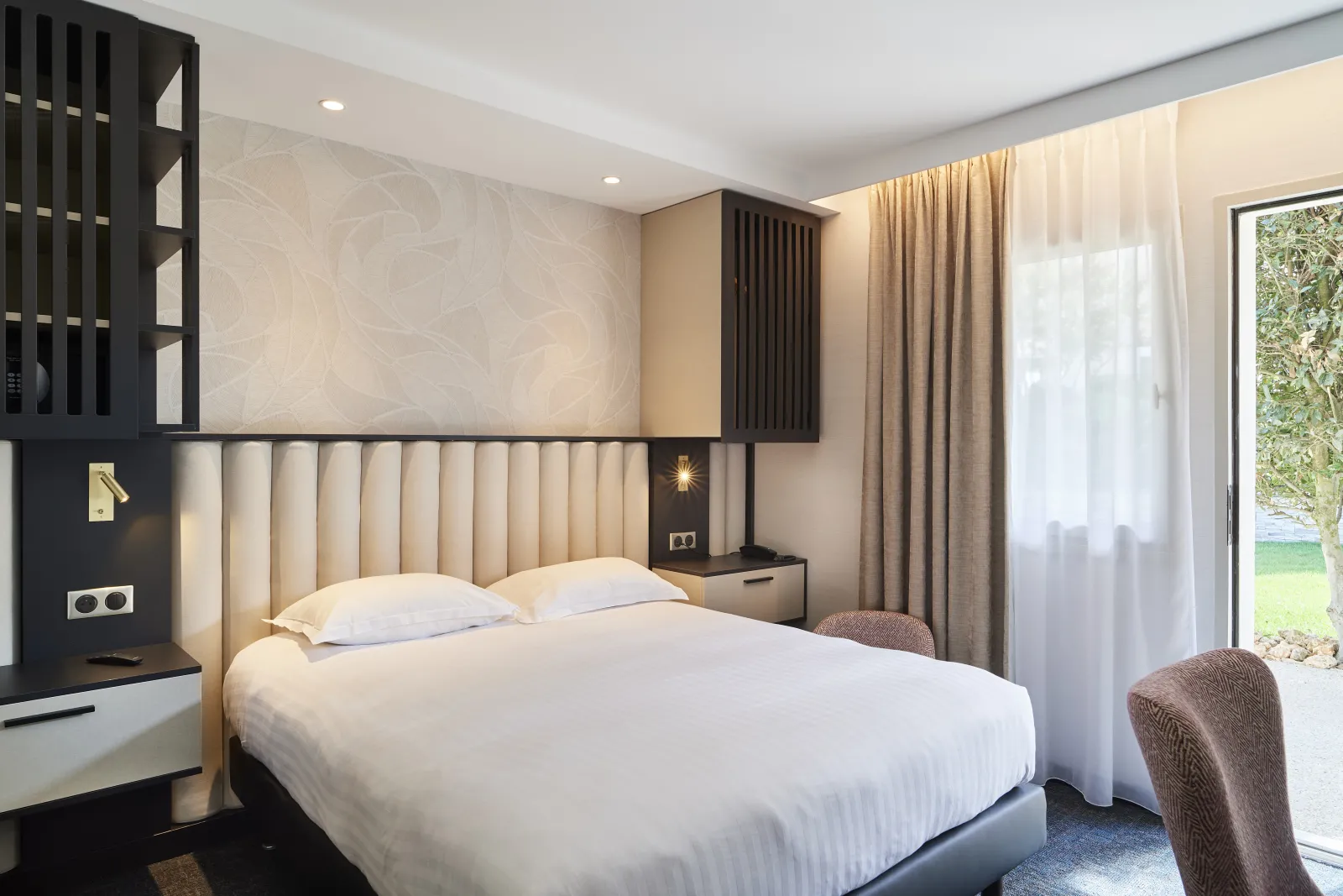 Modern hotel room featuring a double bed with white linens, beige upholstered headboard, black and wood furnishings, large window overlooking garden, beige curtains and warm ambient lighting.